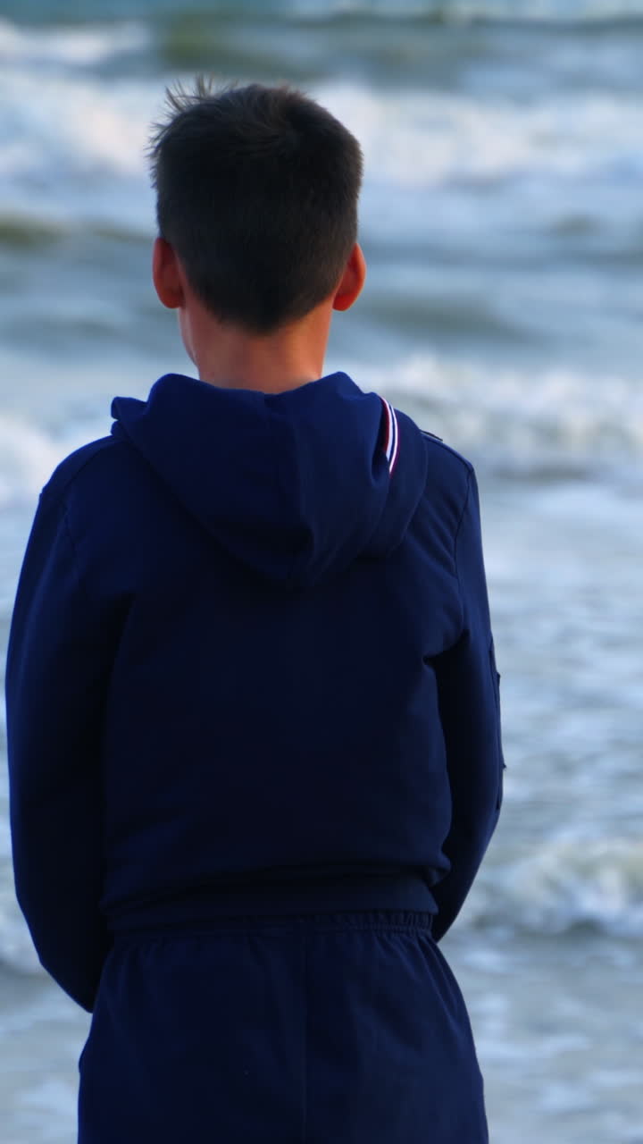 Back view of a boy near the blue sea. Teenage boy standing on the beach and looking on the sea water. Child dreaming on the ocean shore. Vertical video