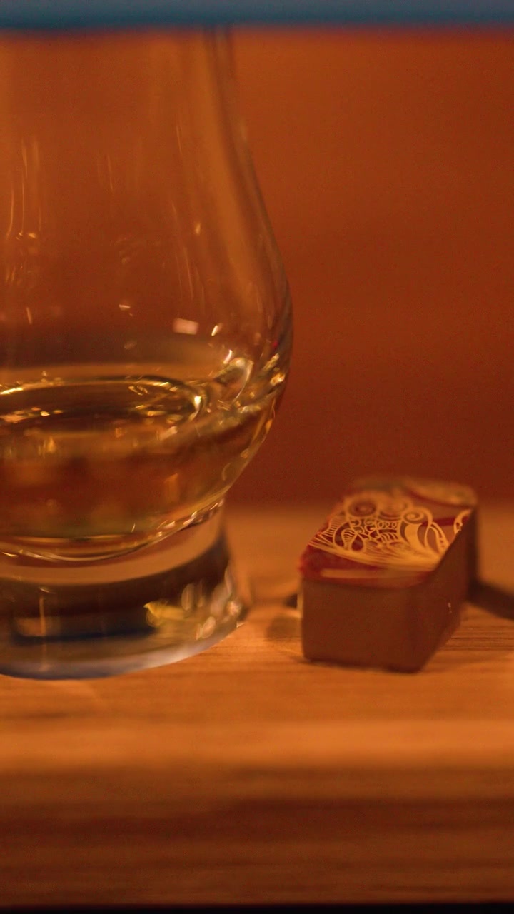Lenta panoramica della telecamera rivela un bicchiere di whisky e cioccolato su un vassoio di legno, illuminazione ambientale calda.