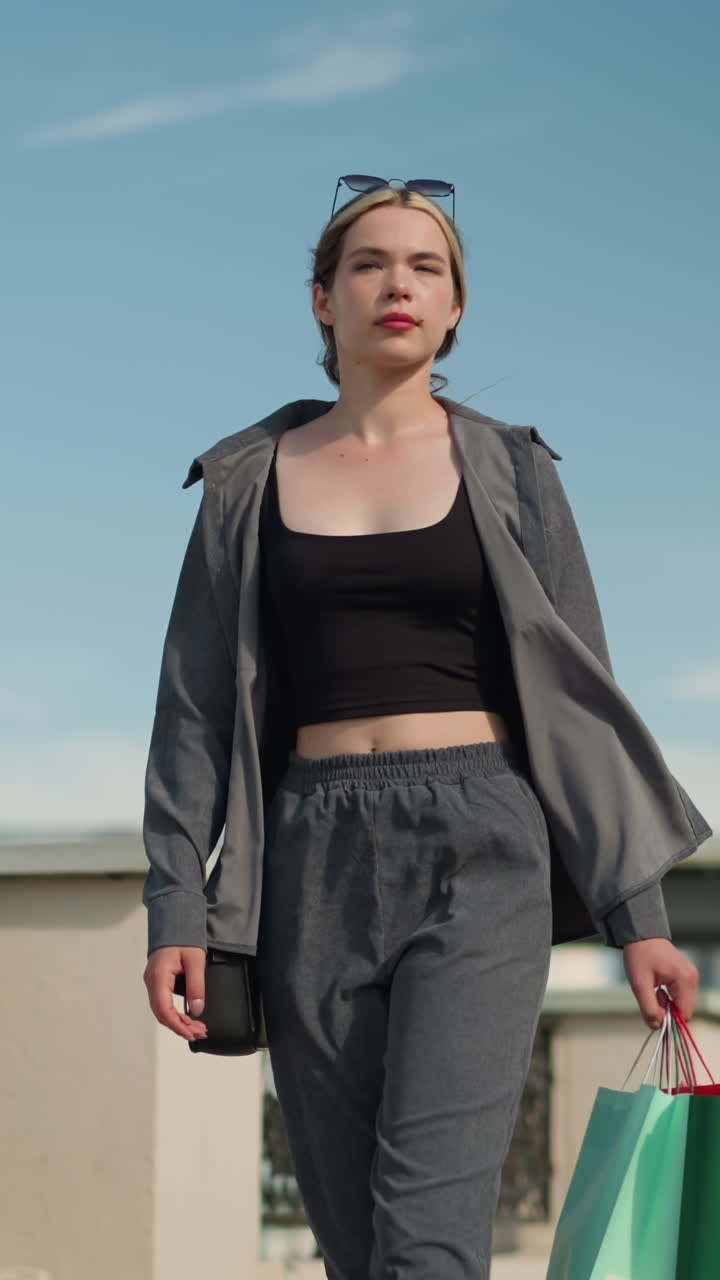 dama blanca en un traje gris casual caminando cerca de una valla del puente, sosteniendo tres bolsas de compras de colores, mirando hacia la derecha. el fondo muestra coches pasando por el puente superior con un cielo azul claro