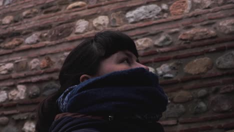 close shot of a brunette young girl in winter clothes standing next to a maroon brick wall of an old building, looking upwards