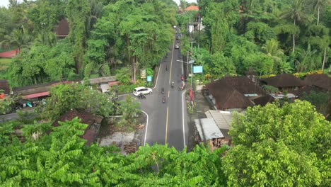 drone shot revealing lush dense tropical trees and busy intersection road with motorcyclists and cars passing at high speed in traditional balinese district