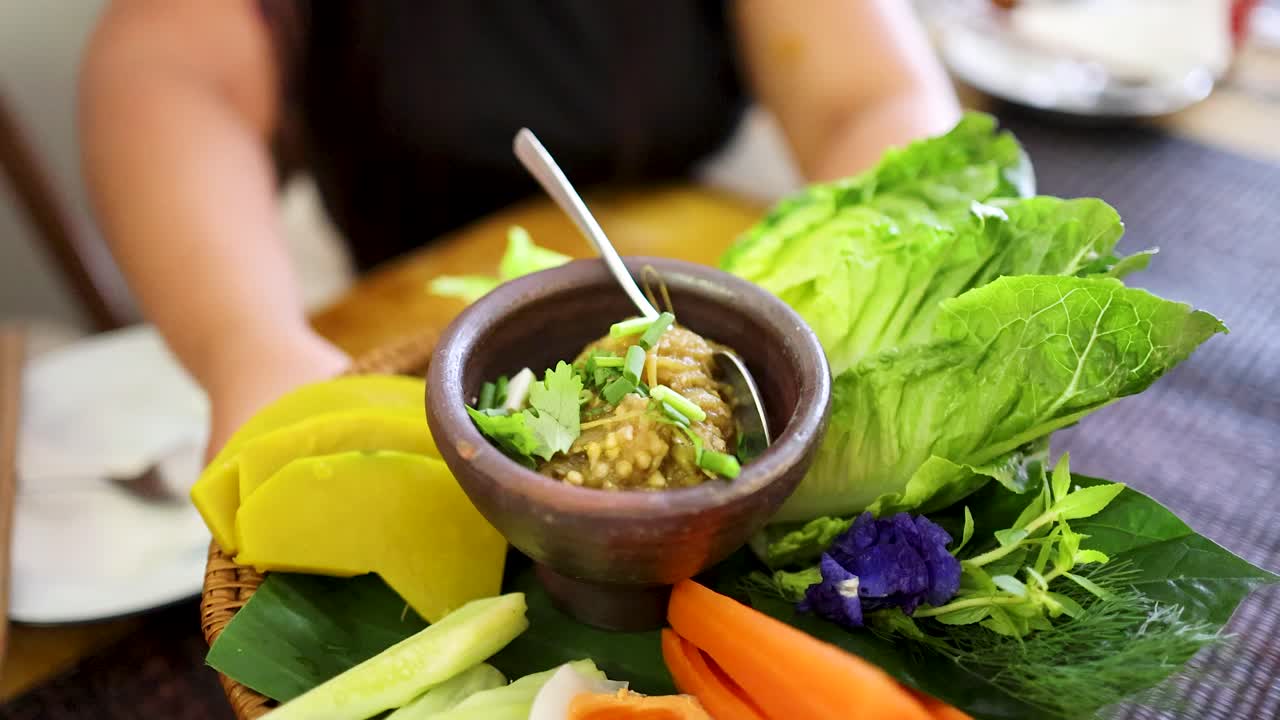 A person presents a bamboo basket of roasted green chili dip and assorted fresh vegetables in bright natural light, highlighting Northern Thai cuisine and vibrant colors