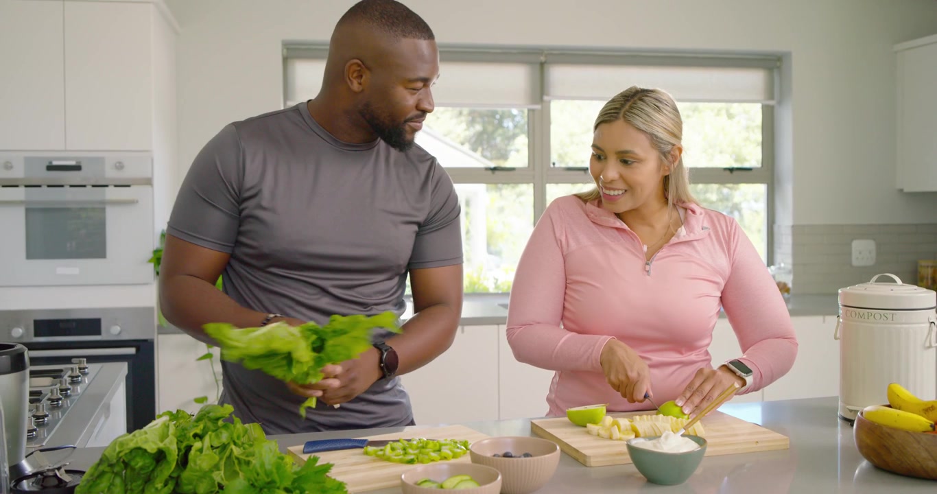 Casal preparando salada na ilha da cozinha, picando alface, cortando frutas em cubos, arrumando a alface e sorrindo.