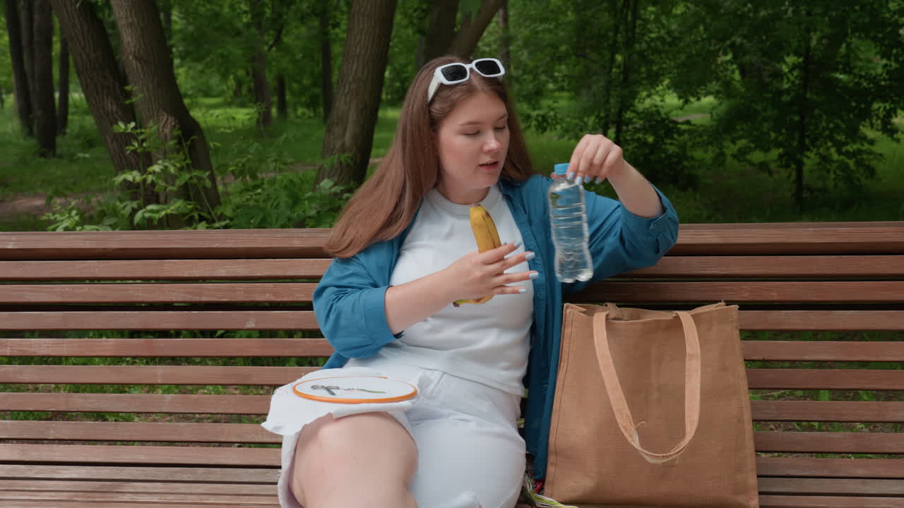 Young lady sitting on wooden bench in green park with fabric and scissors on lap takes out ripe plantain and bottle of water from bag, relaxing outdoors surrounded by trees under natural daylight