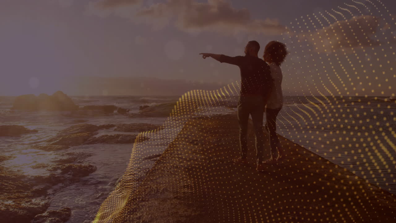 Pointing towards ocean, couple standing on rocks with digital wave animation over scene