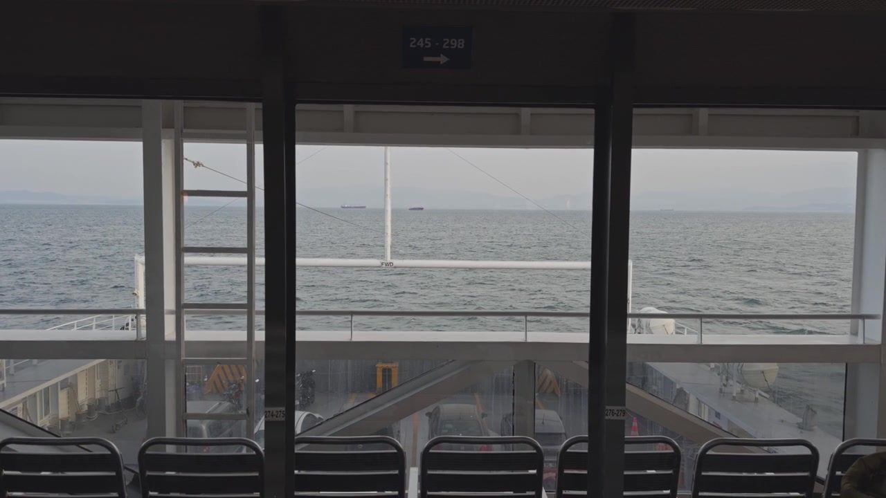 Inquadratura statica dall'interno di un traghetto per auto, che guarda attraverso i finestrini della cabina il mare aperto, con il ponte veicoli sottostante e sedili passeggeri vuoti in una giornata fosca.