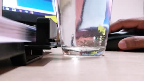 Eine-Hand,-Die-Mit-Einem-PC-Auf-Einem-Schreibtisch-Arbeitet,-Wobei-Der-Schwerpunkt-Auf-Dem-Vollen-Wasserglas-Liegt