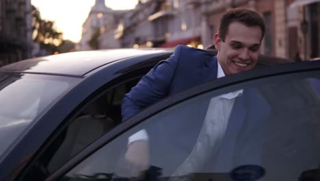 Un-Hombre-Guapo-Y-Confiado-Con-Ropa-Formal-Azul-Sale-Del-Auto.-Un-Joven-Empresario-Abre-La-Puerta-Del-Auto-Y-Mira-La-Cámara,-Sonriendo