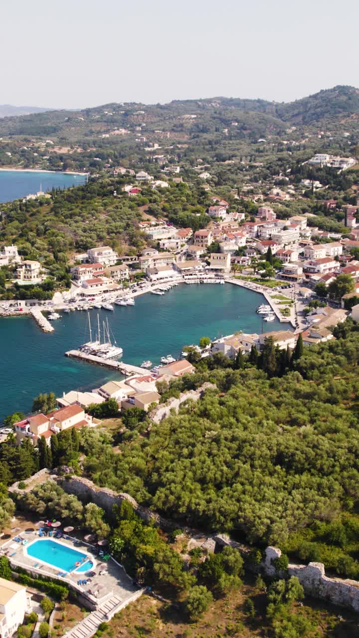 Aerial drone view of the quaint coastal village of Kassiopi in Corfu, showcasing its traditional houses, marina, and turquoise Ionian shoreline