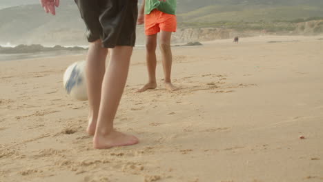 Two-faceless-Brazilian-kids-playing-soccer-barefoot-at-the-beach,-unrecognizable-boys-kicking-soccer-ball