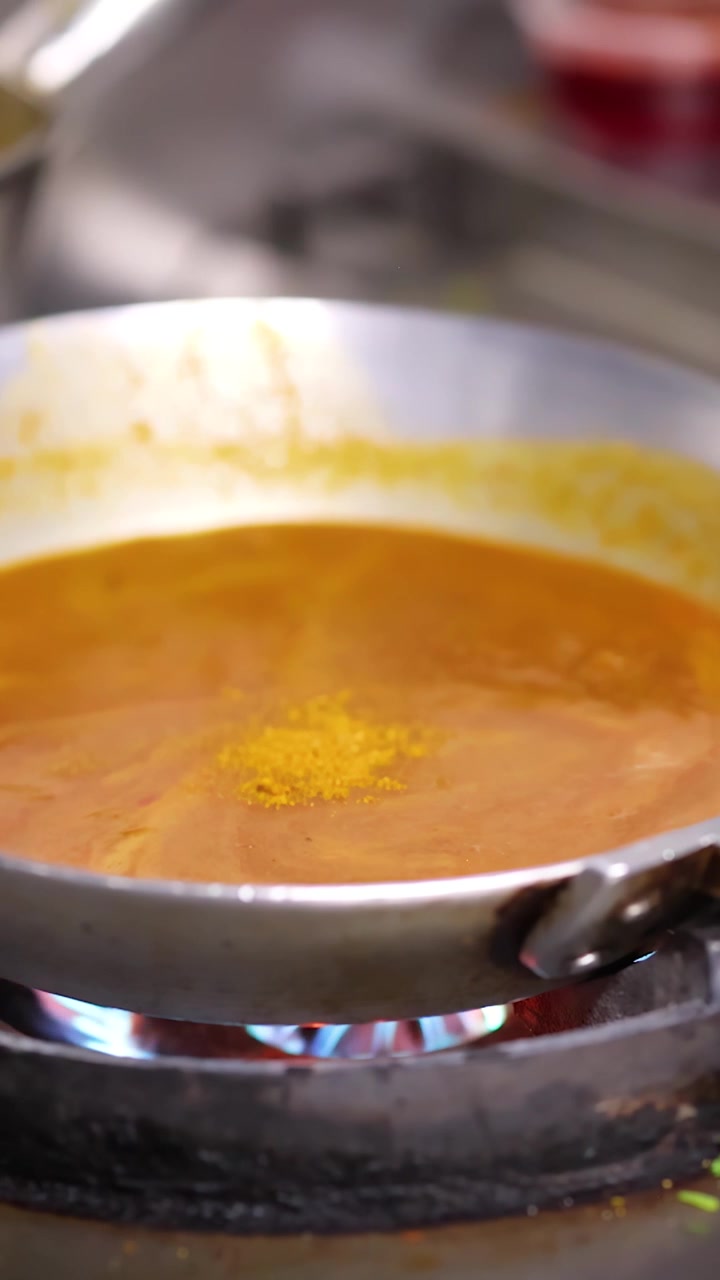 Primo piano di polvere di spezie gialla che cade in salsa al curry arancione che sobbolle su fornello a gas.
