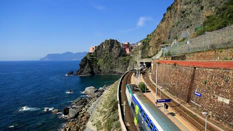 static view of stunning train station, railway and tracks alongside beautiful rocky italian coastline and clear blue ocean water while train drives through on sunny summer day