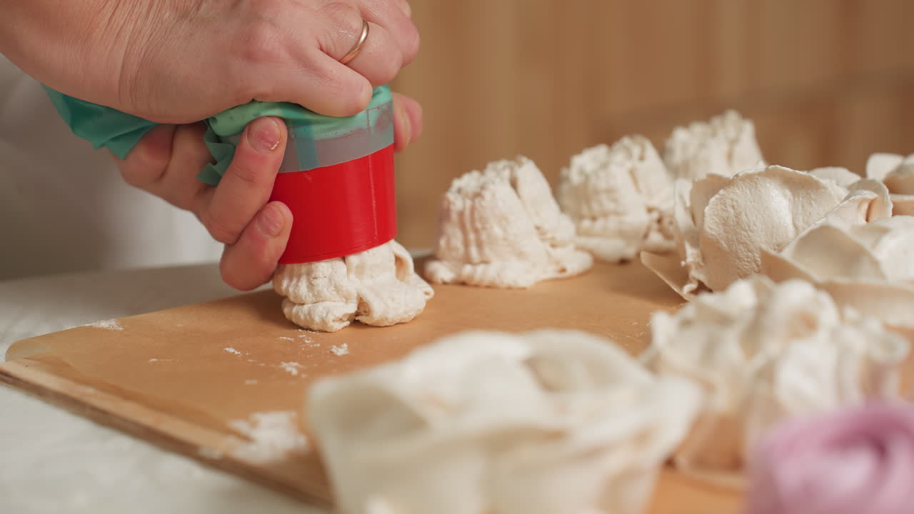 Close up of caterer pressing piping bag with red nozzle as creamy dough drops onto parchment paper, forming delicate swirl designs for baking preparation in bright kitchen environment
