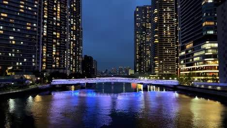 colorful lights illuminate a city bridge at night reflecting in the calm river water