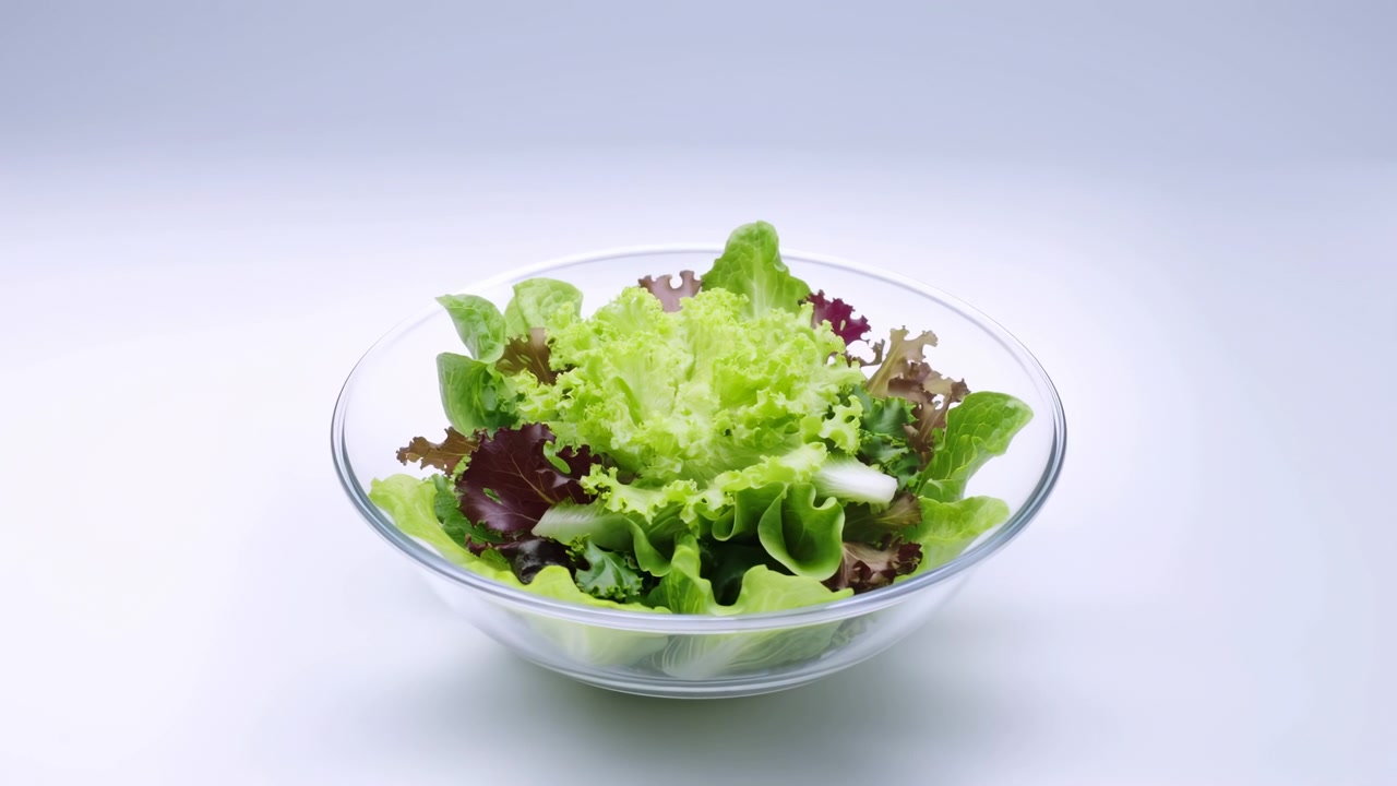 Blandet salat arrangert i en klar glassbolle på en hvit bordplate, med krusete lysegrønne blader, dype burgunderfargede aksenter, myk, kjølig gradientbakgrunn, frisk tekstur og en ren, lys presentasjon.