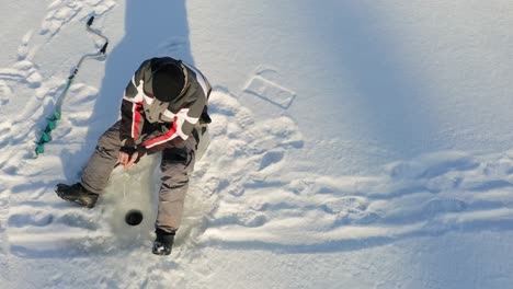 man sitting on the snow and fishing from a small hole on a sunny winter day