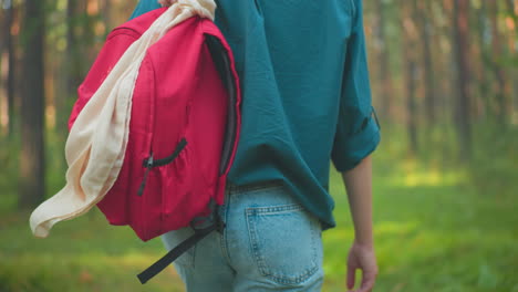 vista trasera de una mujer caminando por un bosque iluminado por el sol con una mochila roja colgada sobre su hombro, ajustando la bolsa con una mano, lleva una camisa verde, mezclándose con el entorno natural