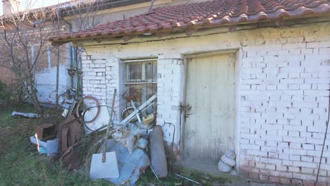 Cobertizo-De-Ladrillo-Encalado-Con-Techo-De-Pizarra-Roja-Y-Una-Puerta-De-Madera-Con-Algunos-Trastos-Apilados-Delante