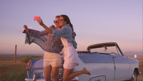 Hermosas-Amigas-Tomando-Selfies-En-Un-Viaje-Por-Carretera-Al-Atardecer-Con-Un-Coche-Antiguo