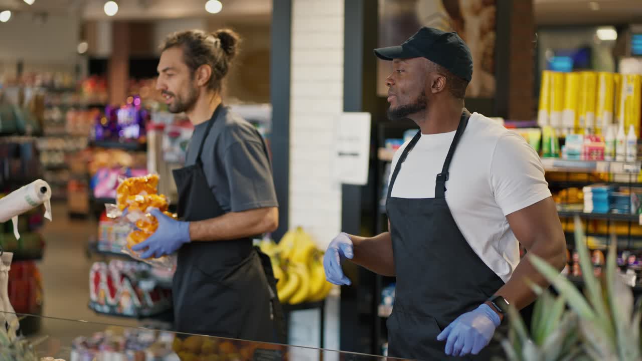 A man with Black skin in a white T-shirt and a Black apron tells his colleague a man with a beard in a gray T-shirt where to put certain products during their work together in a large supermarket