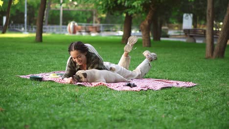 Woman-with-dreadlocks-playing-with-her-dog-on-grass,-laying-on-plaid---tricking-him-with-a-little-snack