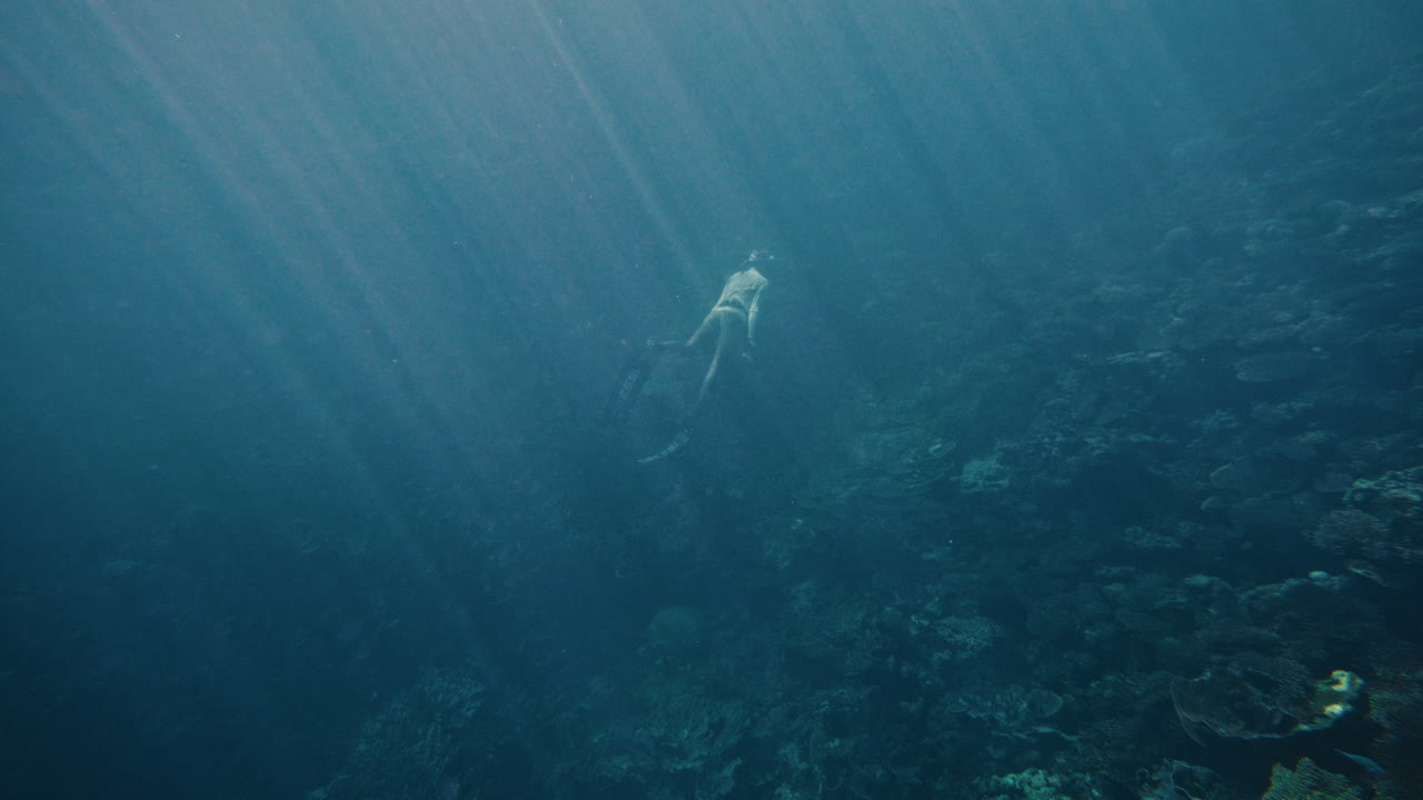 Free diver slowly kicks long blade fins as sun beam light rays shine down underwater in slow motion