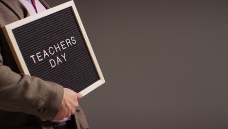 close up studio shot of male teacher standing against grey background holding notice board reading teachers day 1