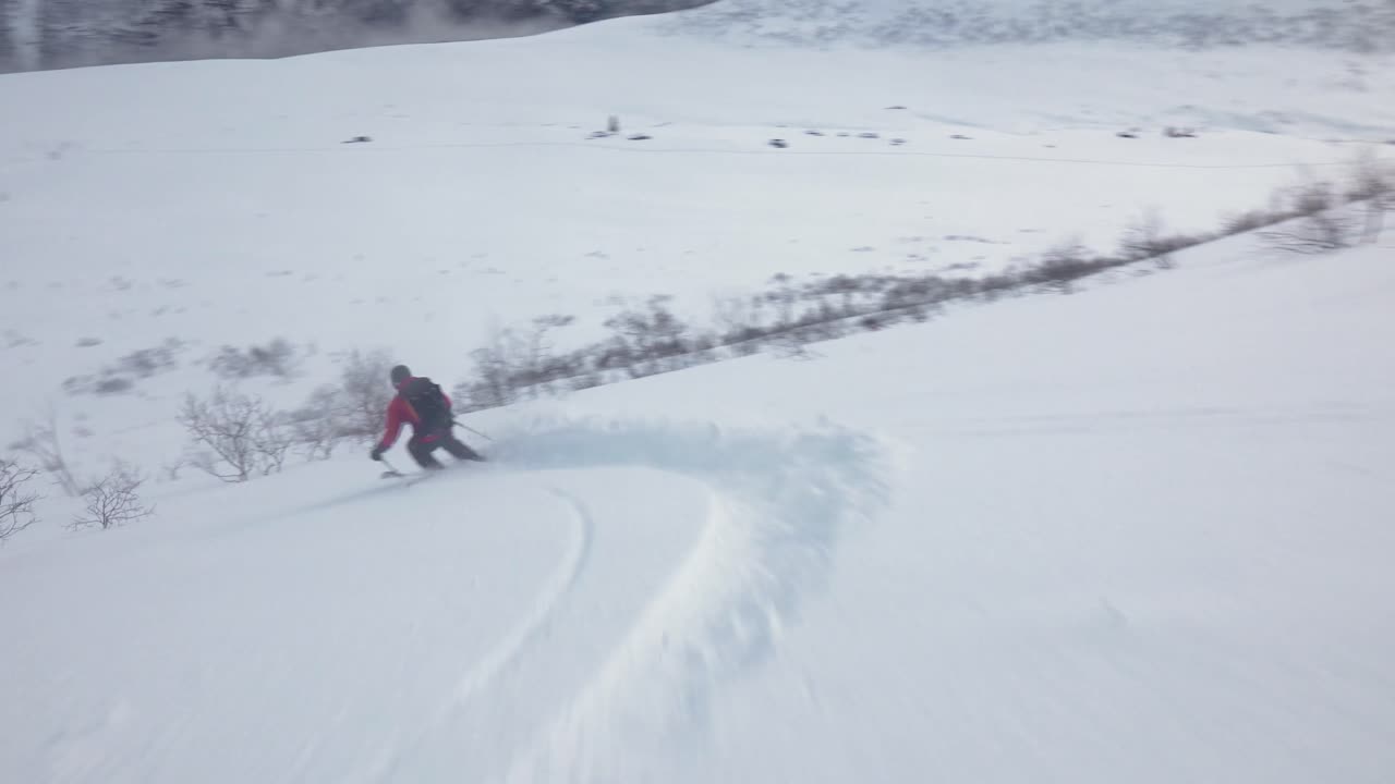 hombre con chaqueta roja esquiando cuesta abajo, zigzagueando a través de pequeños arbustos con hermosos paisajes en el fondo