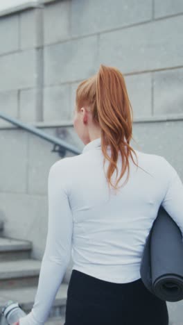 a young woman in activewear carrying a yoga mat and water bottle while leaving a workout session in an urban environment