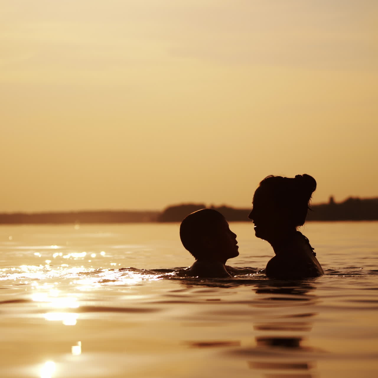 Mother and boy inside the river water at sunset. Silhouette of a woman throwing a boy up into the evening water. Happy family holidays outdoors.