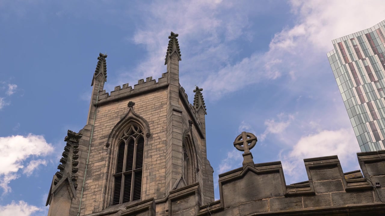 Inquadratura dal basso della torre in pietra e della croce della Chiesa di San Giovanni Evangelista, stagliate contro nuvole alla deriva con un moderno grattacielo