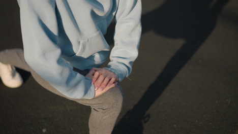 partial view of lady in blue sweater exercising outdoors, performing a kneeling stretch with shadow cast on tarred ground under bright sunlight