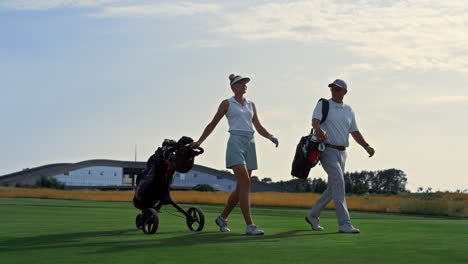 Two-golfers-walk-fairway-at-sunset.-Rich-couple-carry-sport-equipment-clubs.