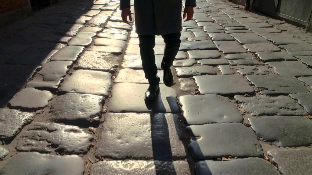 Uomo che cammina con un cappotto lungo mentre inizia a fare un passo verso l'obiettivo posizionato in basso su una strada acciottolata storica, ombra lunga