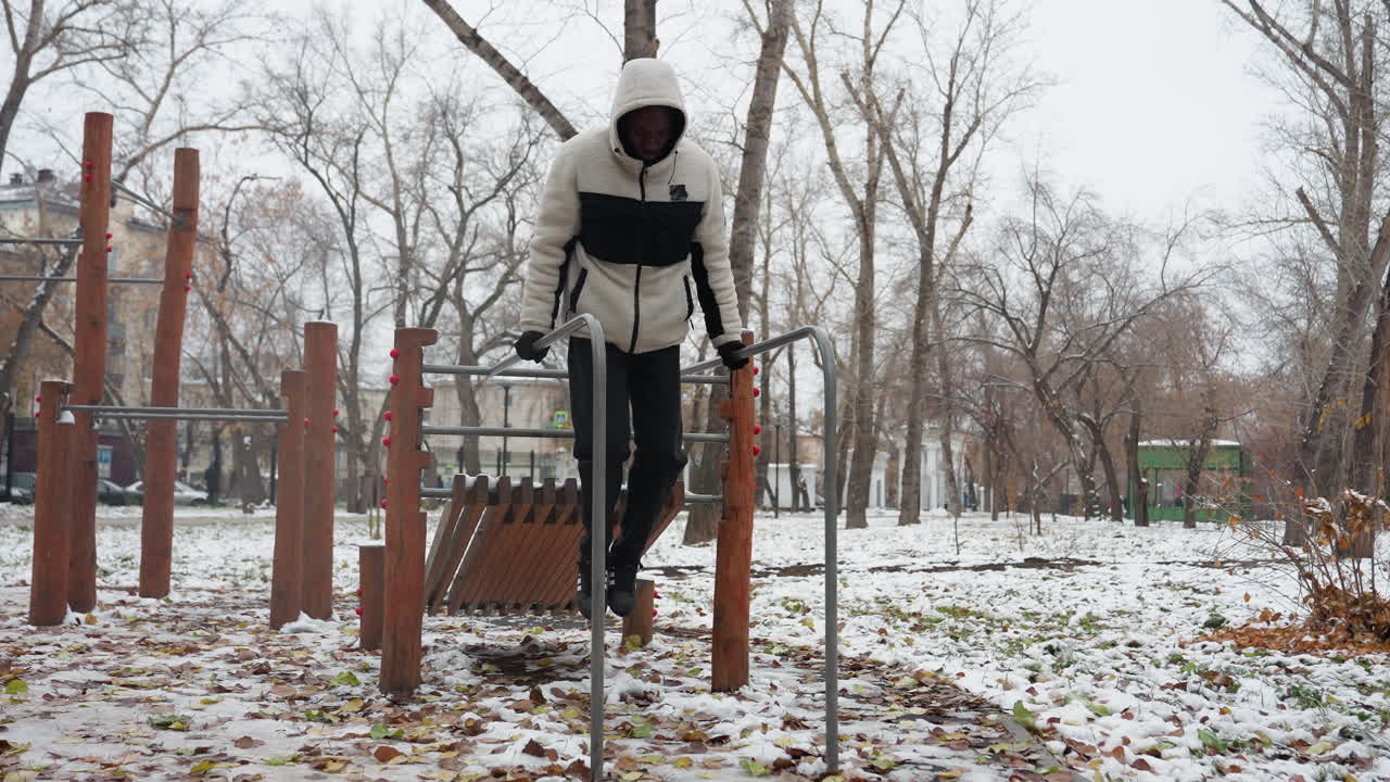 젊은 성인은 눈 인 야외 공원에서 평행 바에서 근육 구축 운동을 수행합니다. 따뜻한 겨울 옷을 입고 차가운 환경에서 운동하는 동안 균형과 힘을 유지합니다.