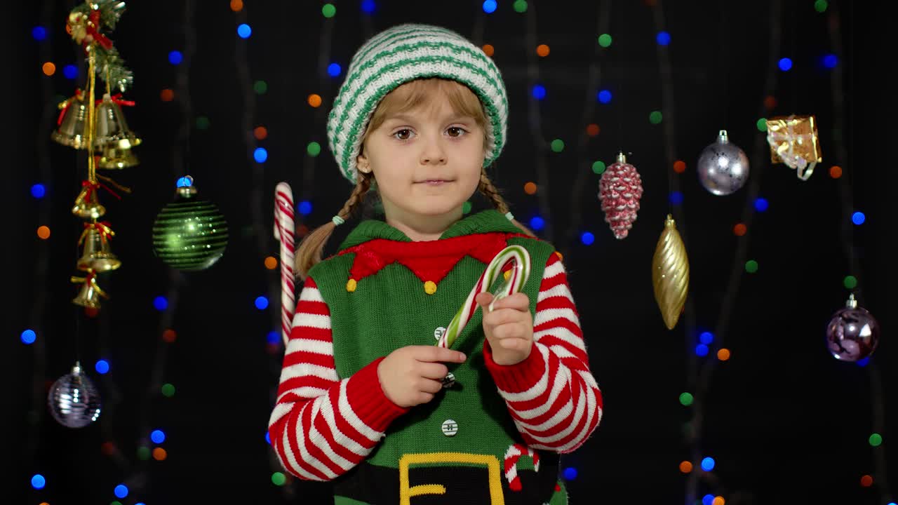 Malá holčička oblečená jako elfka se usmívá a drží cukrovou hůlku.