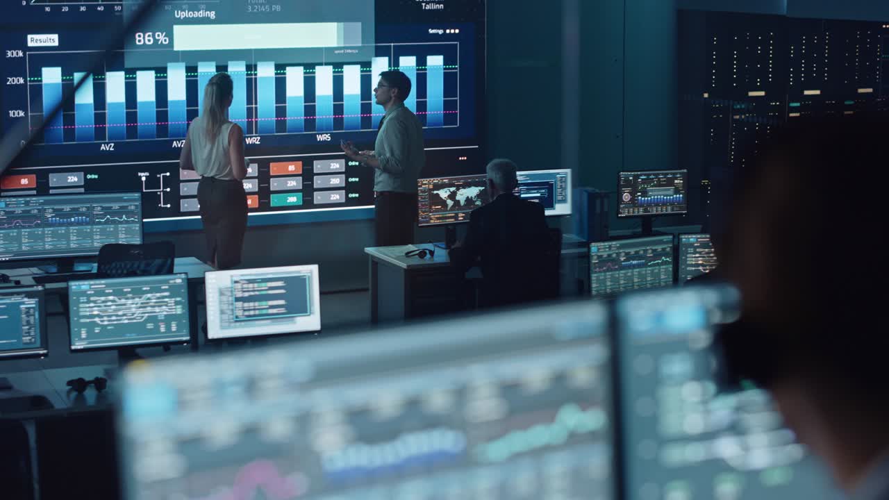 dos ingenieros de software hablando junto a una gran pantalla en una oficina de monitoreo moderna con alimentación de análisis en vivo con gráficos.