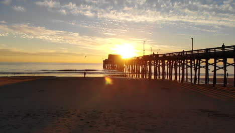 drone flying under the newport beach pier at california sunset with sunlight beams and people in silhouette on the sand aerial