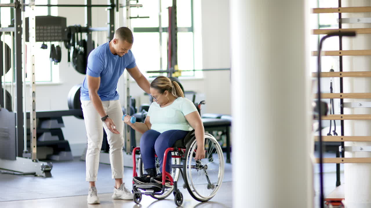 Assisting woman with paraplegia in wheelchair, trainer helping with dumbbell exercise in gym
