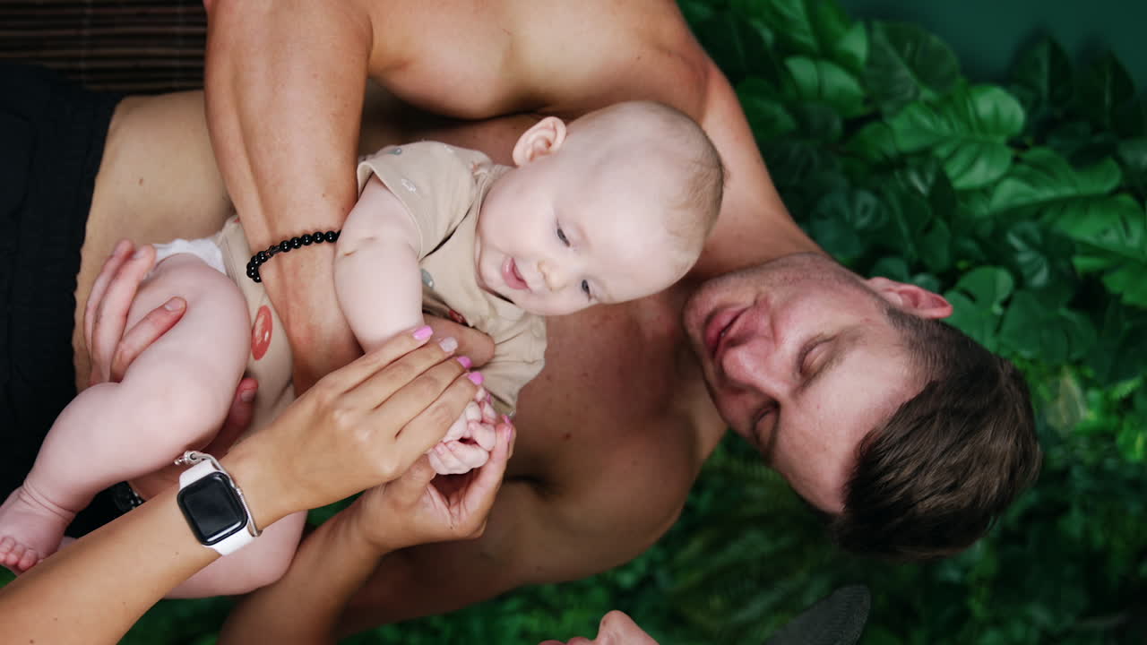 Caucasian dad holding a baby boy in front of him. Female hands clap tiny kid's hands. Vertical view.