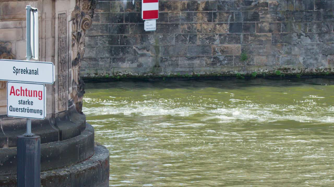 Fixed camera captures warning sign and strong water current by stone wall in Berlin canal