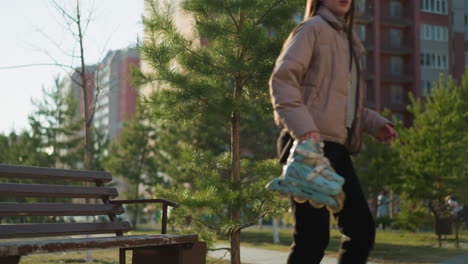 a girl sits on a park bench, wearing a peach jacket, grey inner shirt, and black trousers. she picks up her rollerblades and prepares to leave, creating a serene and reflective moment in the park