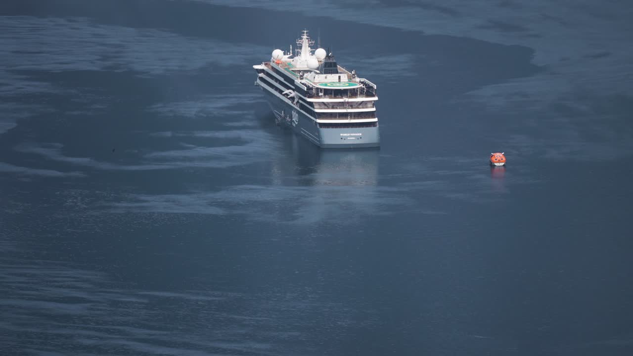 A large cruise ship is anchored in the calm waters of Geiranger Fjord, its reflection shimmering on the water's surface. Motorboats cruise nearby.