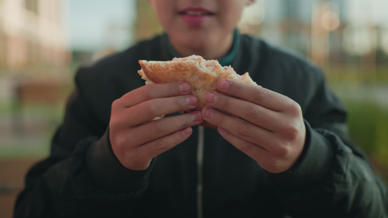 Close up boy savoring pizza with both hands, focus on bread texture and enjoyment of bite, outdoor casual moment in warm daylight with blurred background