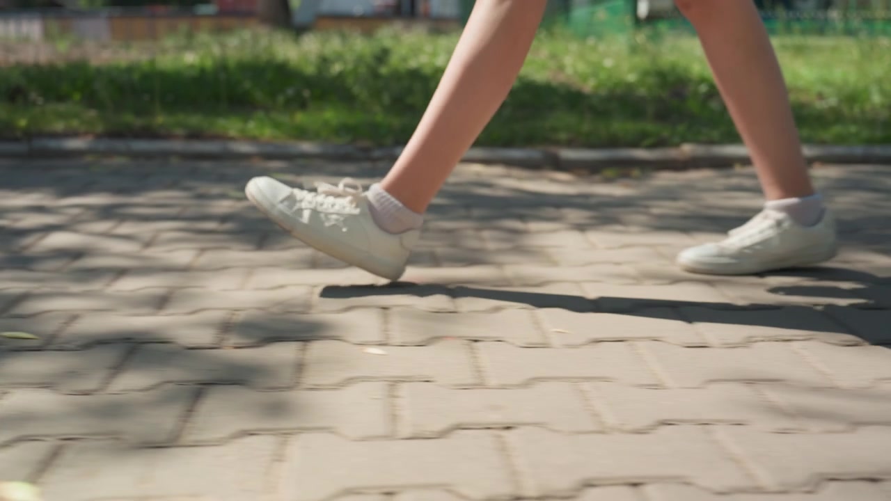 Beyaz spor ayakkabı giyen bir kişinin bacakları ve ayaklarının yakın çekimi, çimenli arka plana sahip parke zeminde yürüyor. Ayak hareketi, dış mekanda hareket halindeki alt vücudu yakalayan sürekli adımlamayı gösteriyor.