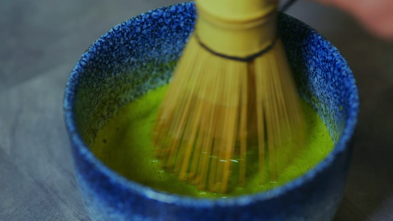 A close-up shot of a bamboo whisk stirring vibrant green matcha in a textured blue bowl. The motion creates a smooth, frothy blend, highlighting the traditional Japanese tea preparation process