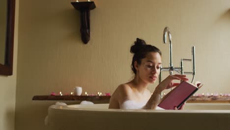 mujer biracial relajada acostada en el baño con espuma y leyendo un libro