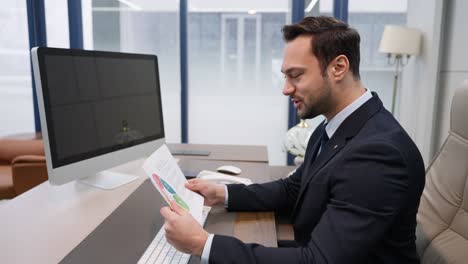 a satisfied happy businessman in a suit analyzes colorful successful performance charts and graphs while seated at his desk with a computer in a modern office - slow motion