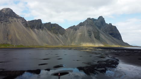 a person walking towards mountains on the black beach with visible mountain reflections in a calm shallow water