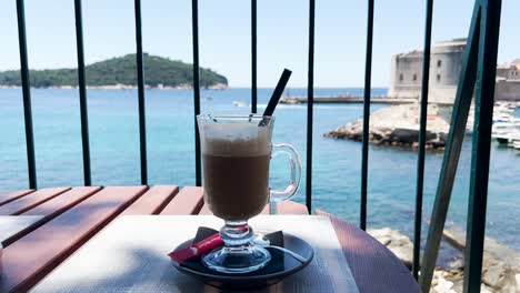 cup of iced latte with a view of blue sea, rocky coast and the old town walls in the background, dubrovnik, croatia